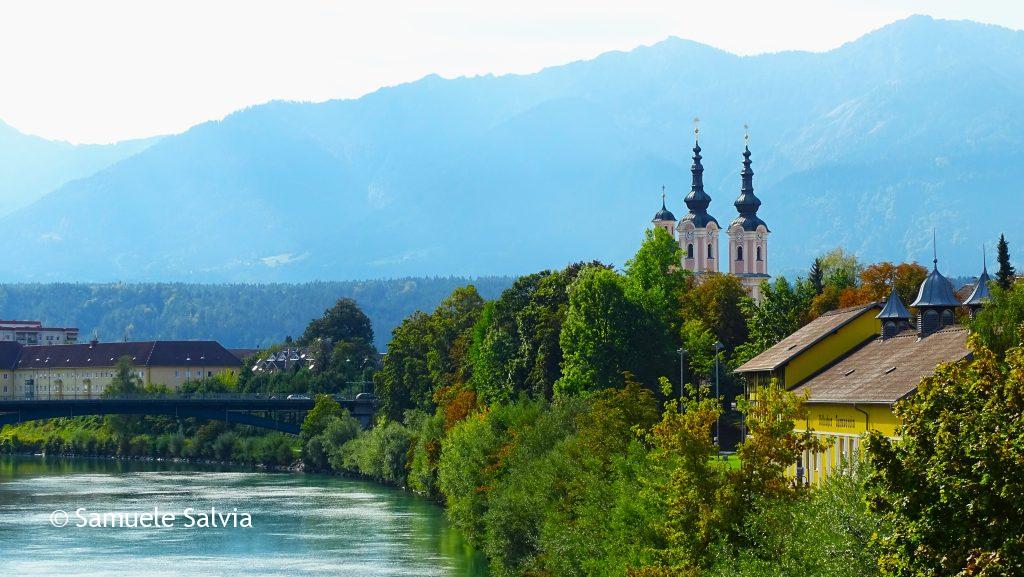 Veduta su Villach dal ponte sulla Drava. Spiccano i campanili della chiesa di Santa Croce, mentre sullo sfondo si ergono le montagne.
